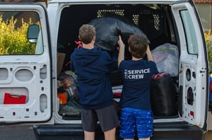 Two men who are taking bags out of the back of a van 